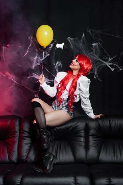 Sexy woman with red hair and clown makeup sitting with yellow balloon on leather couch near spiderweb on black background — Stock Photo