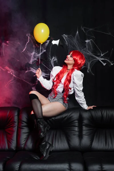 Sexy mujer con el pelo rojo y payaso maquillaje sentado con globo amarillo en sofá de cuero cerca de telaraña sobre fondo negro - foto de stock