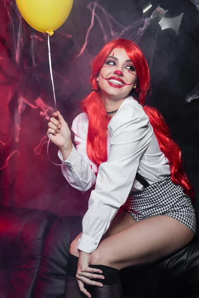 Cheerful woman with red hair and clown makeup holding yellow balloon and looking away on dark background — Stock Photo