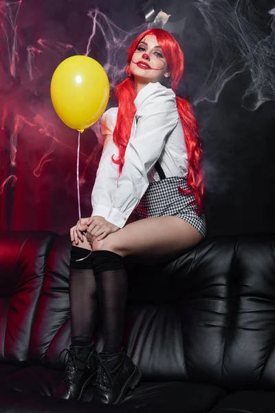 Donna sexy con palloncino giallo e trucco clown seduta vicino ragnatela su sfondo scuro — Foto stock