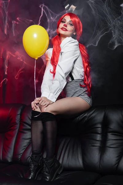 Sexy Frau mit gelbem Ballon und Clown-Make-up sitzt in der Nähe von Spinnennetz auf dunklem Hintergrund — Stockfoto