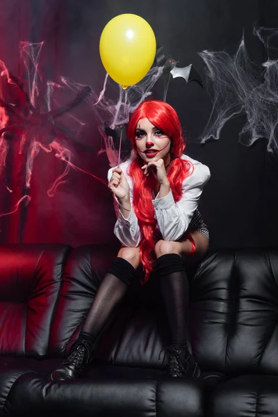 Sexy Frau in Clown-Make-up und schwarzen Kniestrümpfen mit gelbem Luftballon in der Nähe von Spinnennetz auf dunklem Hintergrund — Stockfoto