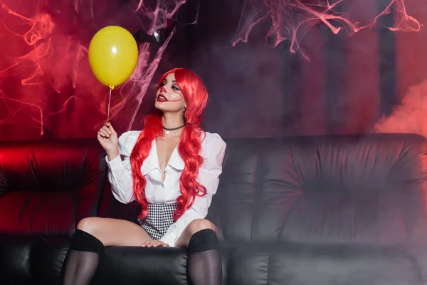 Young redhead woman in clown makeup looking at yellow balloon on dark background with fog and spiderweb — Stock Photo