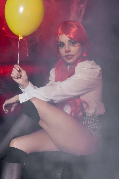 Seductora mujer en maquillaje de payaso sosteniendo globo amarillo en niebla sobre fondo oscuro - foto de stock