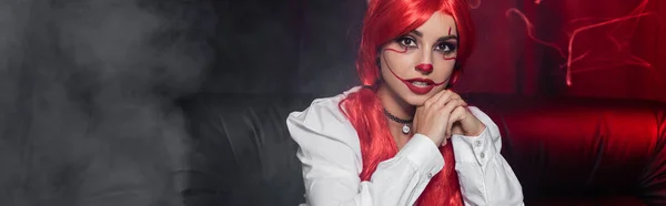 Young woman with red hair and clown halloween makeup looking at camera near fog on dark background, banner — Stock Photo