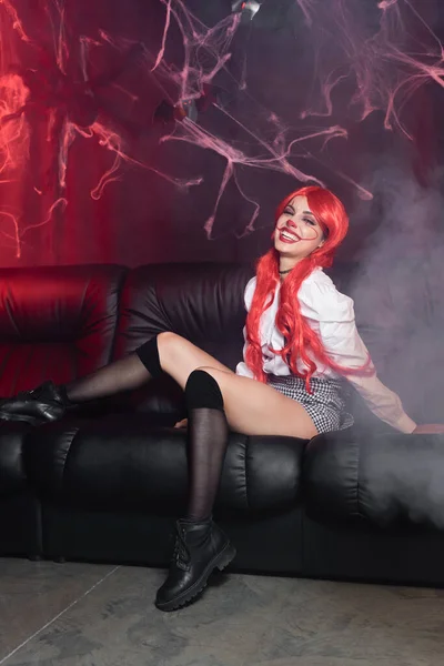 Excited redhead woman in clown makeup and black knee socks sitting on leather couch near spiderweb on dark background — Stock Photo