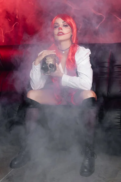 Pelirroja con cráneo dorado y maquillaje de payaso sentada en niebla sobre fondo oscuro - foto de stock