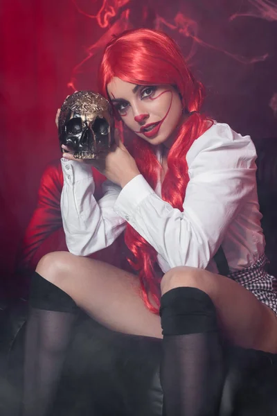 Sexy mujer con el pelo rojo y payaso maquillaje mirando a la cámara cerca de cráneo de oro sobre fondo oscuro con tela de araña — Stock Photo