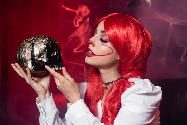 Woman with red hair and clown makeup holding golden skull near spiderweb on dark red background — Stock Photo