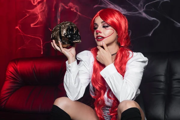 Young redhead woman in halloween makeup looking at golden skull near spiderweb on dark background — Stock Photo