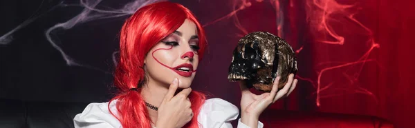 Mujer joven en maquillaje de payaso tocando la barbilla mientras mira el cráneo dorado sobre fondo oscuro con tela de araña, pancarta - foto de stock