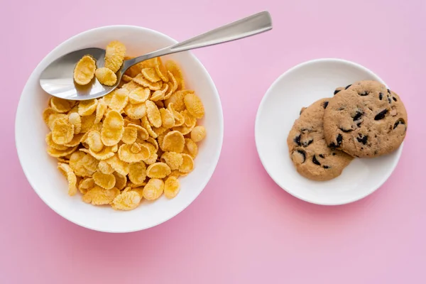 Top view of bowl with tasty corn flakes and spoon near saucer with chocolate chip cookies on pink — Stock Photo