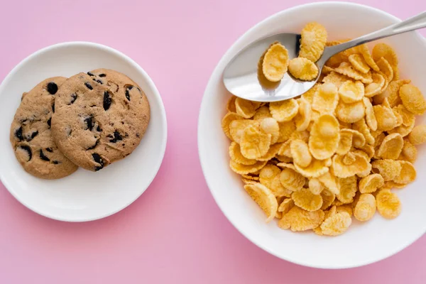 Draufsicht auf Schüssel mit leckeren Cornflakes in der Nähe von Untertasse mit Schokoladenkeksen auf rosa — Stockfoto