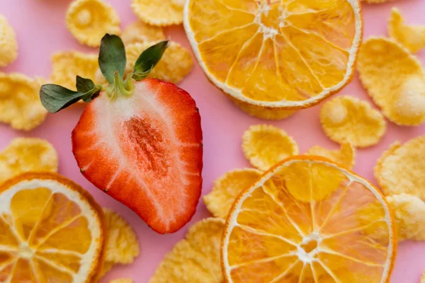Закрыть вид на свежую скользкую клубнику вокруг хрустящих кукурузных хлопьев и сухих апельсинов на розовом — стоковое фото