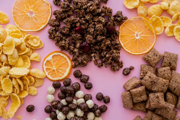 Vista superior de varios cereales de desayuno copos de maíz, soplos, bolas y granola cerca de naranjas secas en rosa - foto de stock