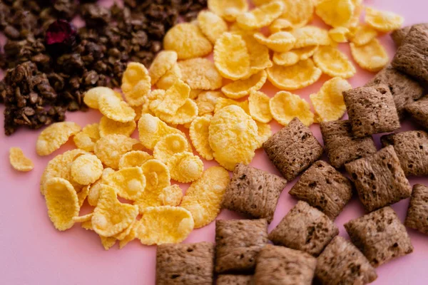 Закрытый вид на разнообразные хлопья кукурузы на завтрак рядом с шейными пуфами и гранолой, изолированной на розовом — стоковое фото