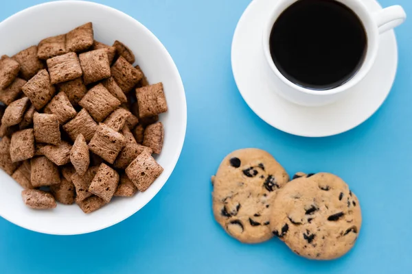 Vista superior de puffs de cereais em tigela branca perto de xícara de café e biscoitos de chocolate chip isolado em azul — Fotografia de Stock