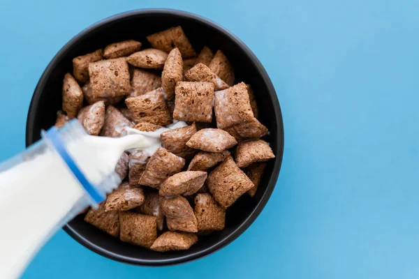 Top view of milk pouring from bottle into bowl with cereal puffs isolated on blue — Stock Photo