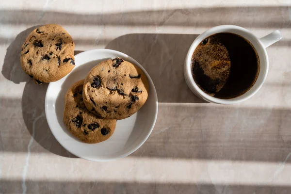 Vue du dessus de la tasse de café noir près de la soucoupe blanche avec biscuits aux pépites de chocolat sur la surface du marbre — Photo de stock