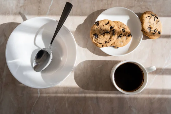 Vue du dessus de tasse de café noir près de biscuits aux pépites de chocolat et soucoupe avec cuillère sur la surface du marbre — Photo de stock