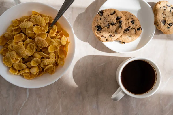Vista superior de la taza de café negro cerca de galletas de chispas de chocolate y tazón con hojuelas de maíz en la superficie de mármol - foto de stock