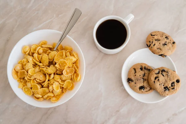 Top view of bowl with corn flakes near cup of coffee and chocolate chip cookies on marble surface — Stock Photo