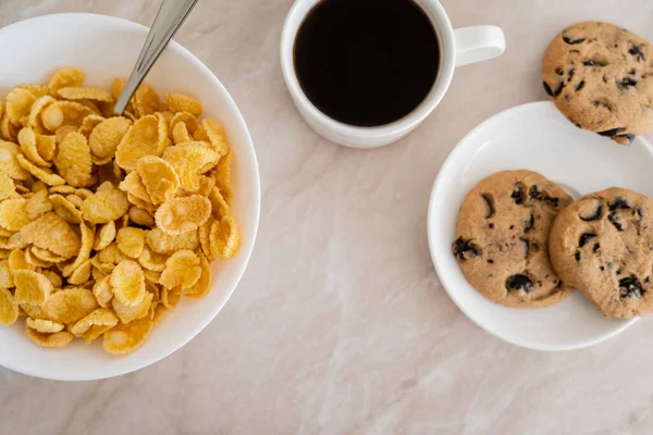 Top view of bowl with corn flakes near chocolate chip cookies and cup of coffee on marble surface — Stock Photo