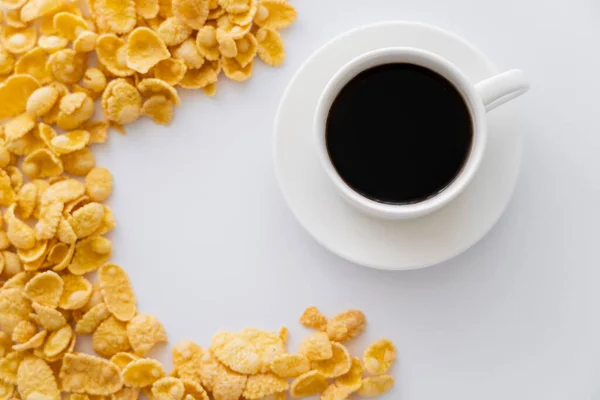 Blick von oben auf süße und knusprige Cornflakes in der Nähe von Tasse schwarzen Kaffee isoliert auf weiß — Stockfoto