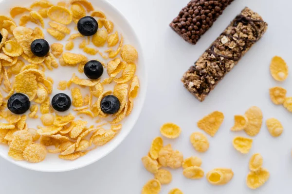 Top view of corn flakes in bowl with organic milk and fresh blueberries near granola bars isolated on white — Stock Photo