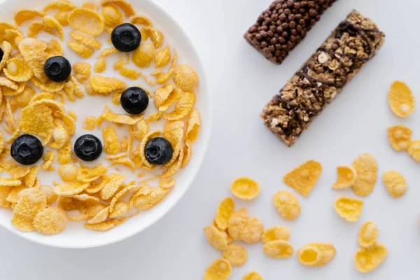 Vista superior de hojuelas de maíz en tazón con leche orgánica y arándanos frescos cerca de barras de granola aisladas en blanco - foto de stock