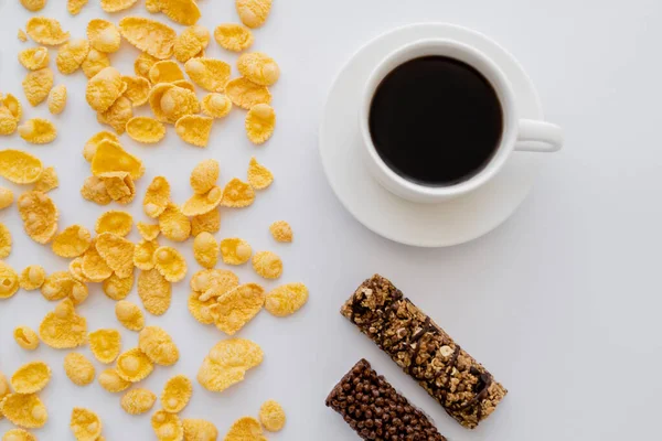 Blick von oben auf knusprige Cornflakes in der Nähe von Tasse schwarzen Kaffees und Müsliriegeln isoliert auf weiß — Stockfoto