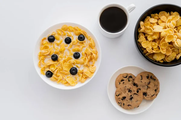 Top view of bowls with corn flakes in milk with blueberries near cup of coffee and cookies isolated on white — Stock Photo