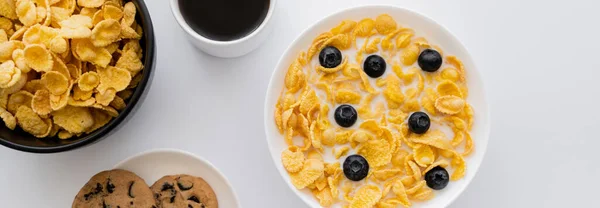 Vue du dessus des bols avec des flocons de maïs dans le lait avec des bleuets près de tasse de café et des biscuits isolés sur blanc, bannière — Photo de stock