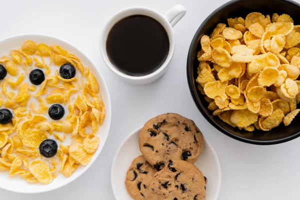 Top view of bowls with corn flakes in milk with blueberries near cup of coffee and chocolate chip cookies isolated on white — Stock Photo