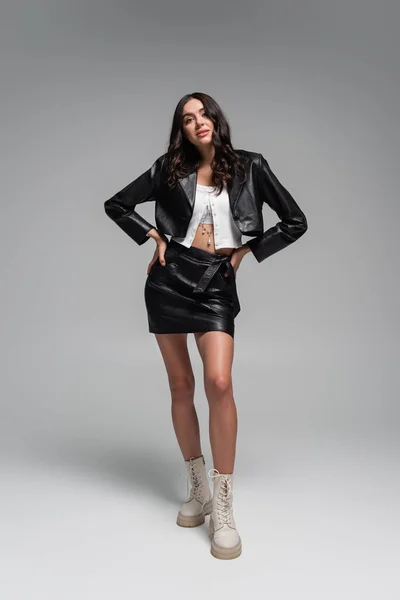 Longitud completa de la mujer joven con estilo en chaqueta de cuero negro y falda posando con las manos en las caderas en gris - foto de stock