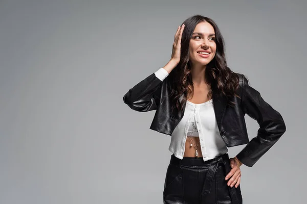 Jovem morena em elegante jaqueta de couro preto sorrindo e ajustando o cabelo isolado no cinza — Fotografia de Stock