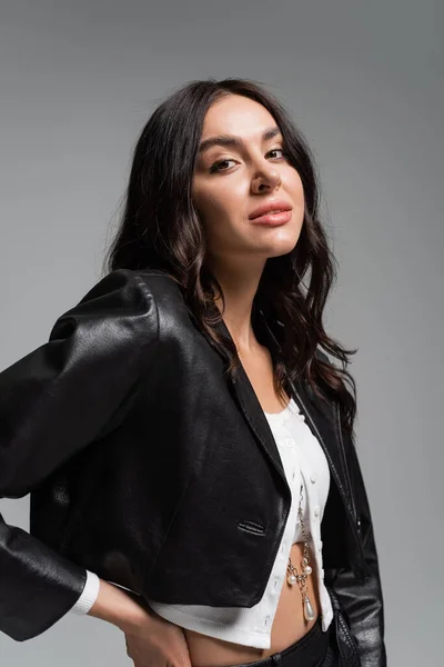 Brunette young woman in stylish black leather jacket standing with hand on hip and looking at camera isolated on grey — Stock Photo