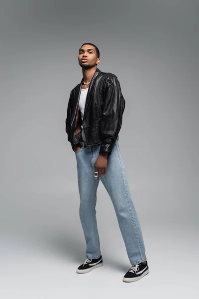 Pleine longueur de jeune homme afro-américain en jeans et veste noire en cuir sur gris — Photo de stock