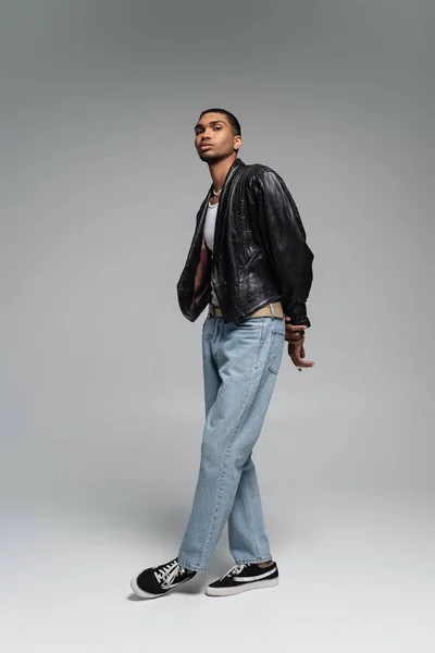 Longitud completa de joven afroamericano hombre en jeans y chaqueta de cuero negro posando en gris - foto de stock