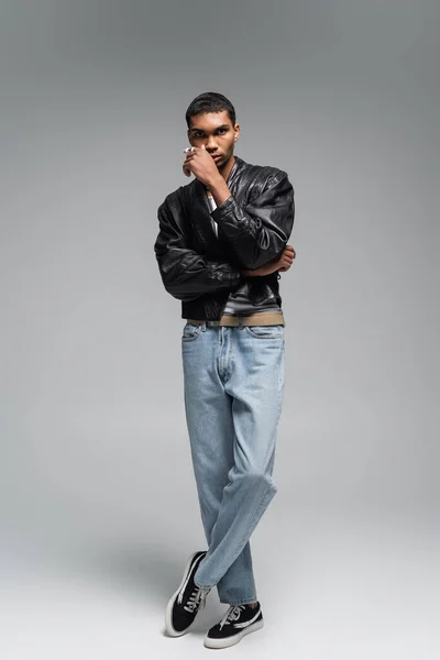 Full length of stylish african american man in jeans and leather jacket posing on grey — Stock Photo