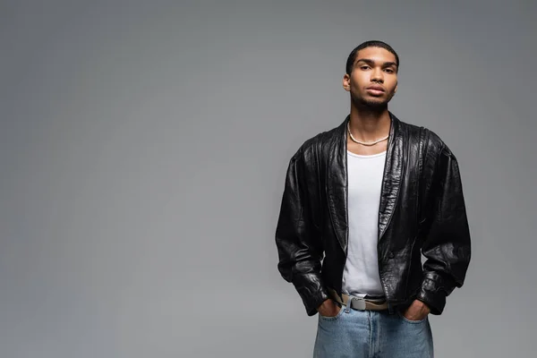Young african american man in jeans and leather jacket posing with hands in pockets isolated on grey — Stock Photo