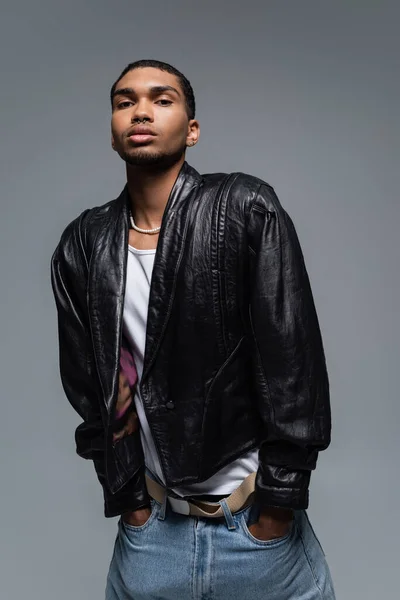 Young african american man in jeans and black jacket posing with hands in pockets isolated on grey — Stock Photo