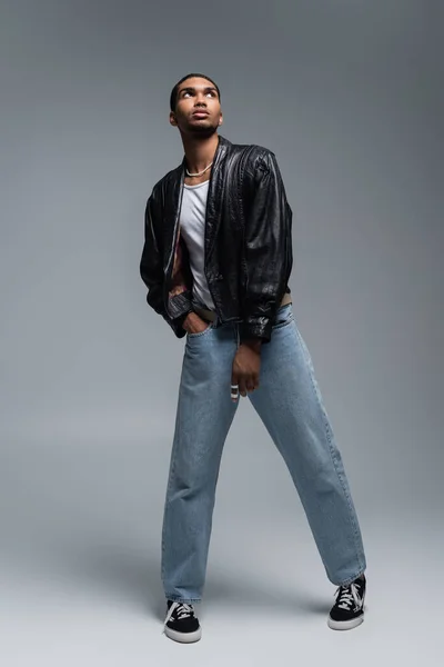 Toute la longueur de l'homme afro-américain élégant en tenue automnale posant avec la main dans la poche de jeans sur gris — Photo de stock