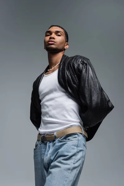 Low angle view of young african american man in jeans and leather jacket posing isolated on grey — Stock Photo