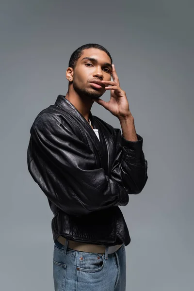 Joven africano americano hombre en cuero chaqueta tocando cara mientras posando aislado en gris - foto de stock