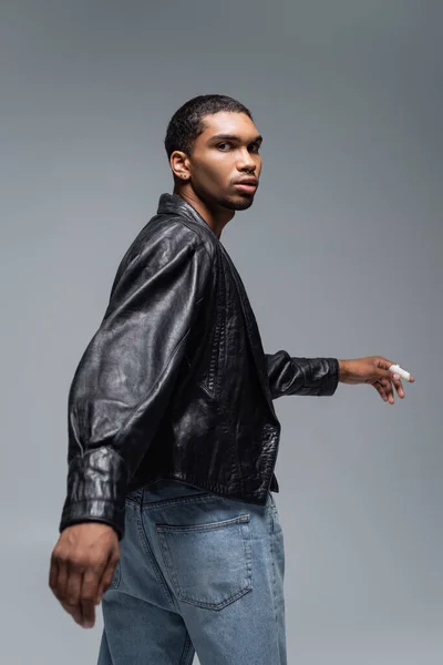 Joven afroamericano hombre en jeans y chaqueta de cuero negro mirando a la cámara aislada en gris - foto de stock