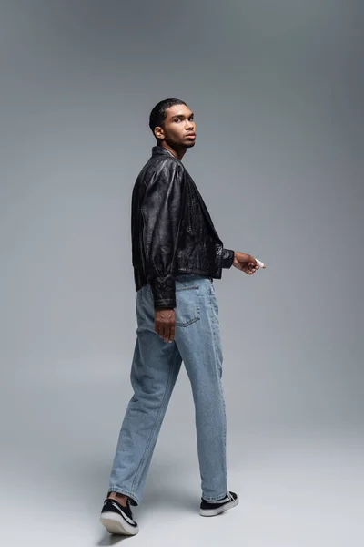 Tutta la lunghezza del giovane afroamericano in giacca di pelle che cammina sul grigio — Foto stock