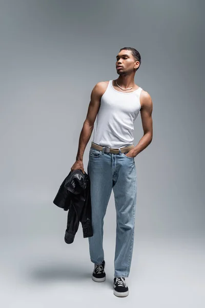 Longitud completa de hombre americano africano muscular en camiseta sin mangas que sostiene la chaqueta de cuero mientras posa con la mano en el bolsillo en gris - foto de stock