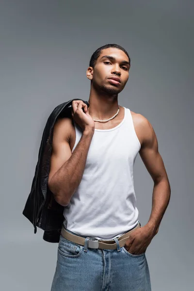 Muscular joven africano americano hombre en tanque superior celebración chaqueta de cuero aislado en gris - foto de stock