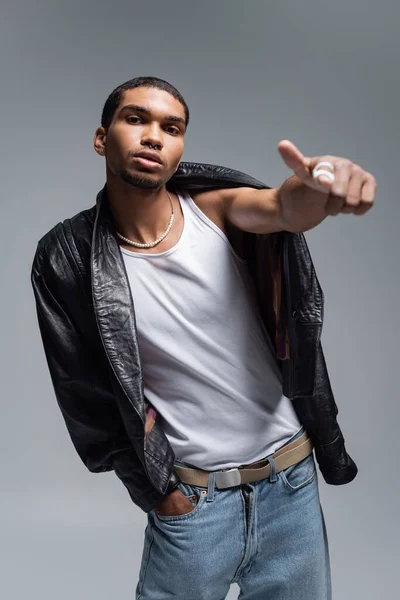 Joven afroamericano hombre en camiseta sin mangas y chaqueta de cuero mostrando el pulgar hacia arriba aislado en gris - foto de stock